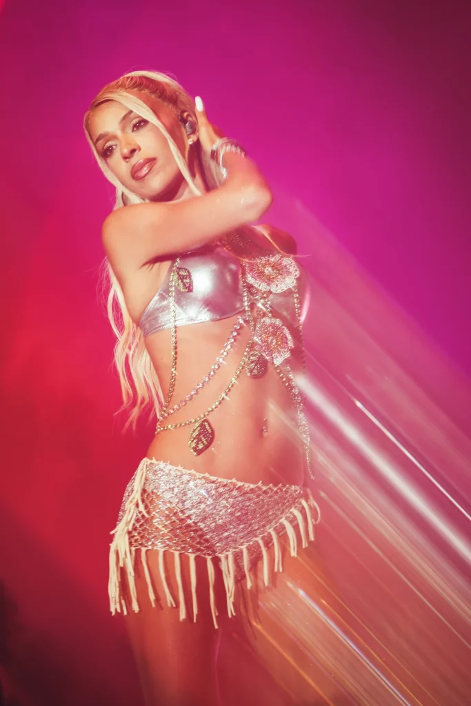 Bad Gyal posando sobre el escenario durante su gira de conciertos, vestida con conjunto plateado y cadenas brillantes, envuelta en iluminación rosa y efectos visuales de espectáculo en directo.