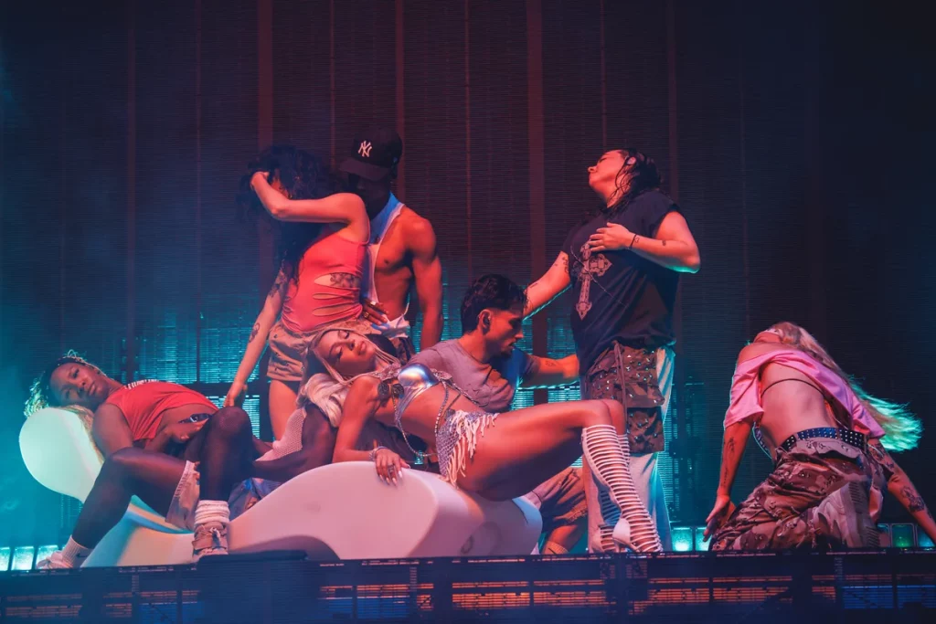 Bad Gyal y su cuerpo de baile realizando una coreografía sobre el escenario durante un concierto en vivo, con iluminación artística y sofá blanco como parte del espectáculo musical.