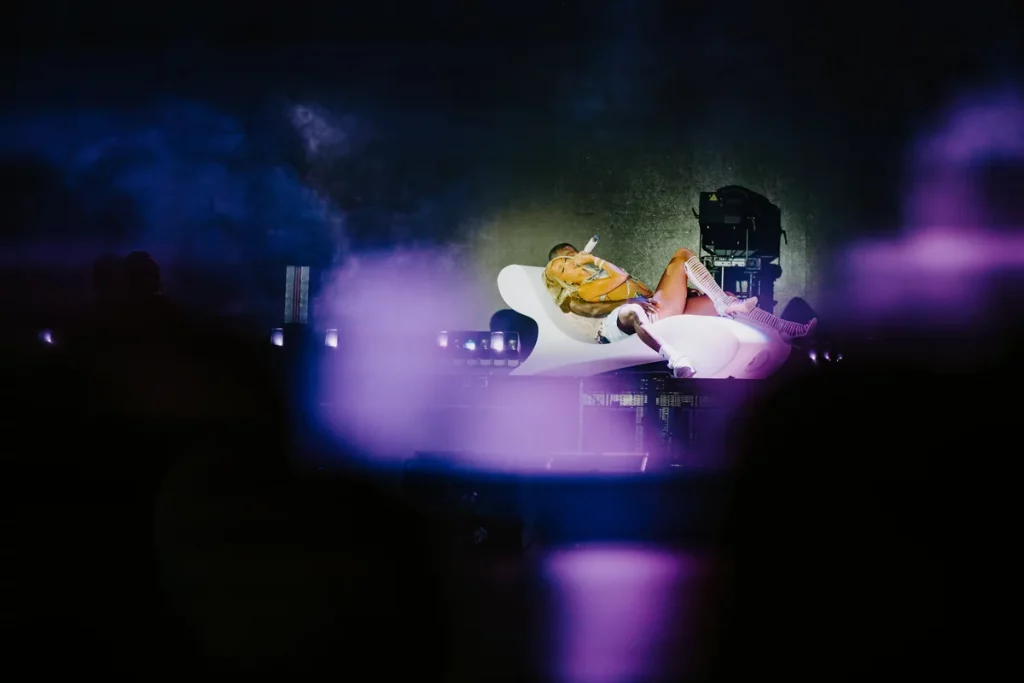 Bad Gyal acostada en un sofá blanco sobre el escenario, rodeada de luces y humo violeta durante su concierto en vivo; fotografía artística de espectáculo musical.