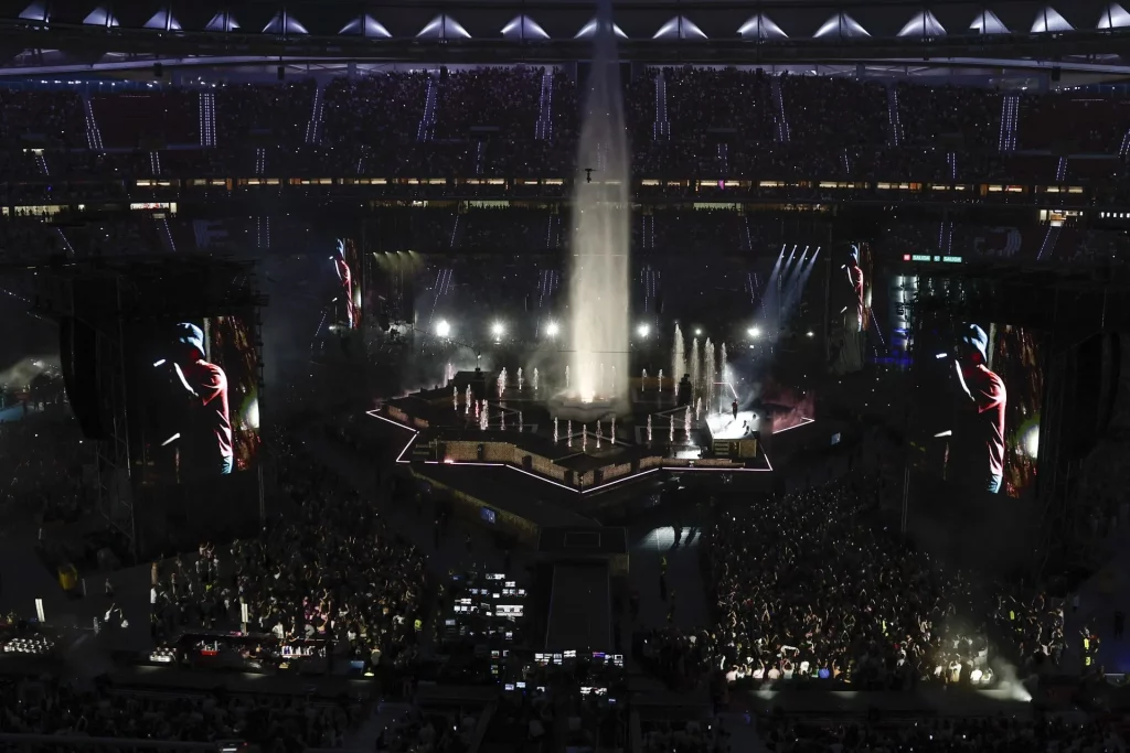Dellafuente en concierto en estadio, actuando sobre escenario central rodeado de fuente de agua y pantallas gigantes, público multitudinario disfrutando del show musical en vivo 2025.