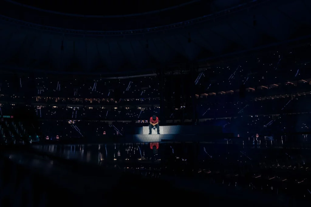 Dellafuente sentado solo en el escenario central de un estadio durante concierto nocturno, rodeado de público y luces, ambiente íntimo en espectáculo musical en vivo.